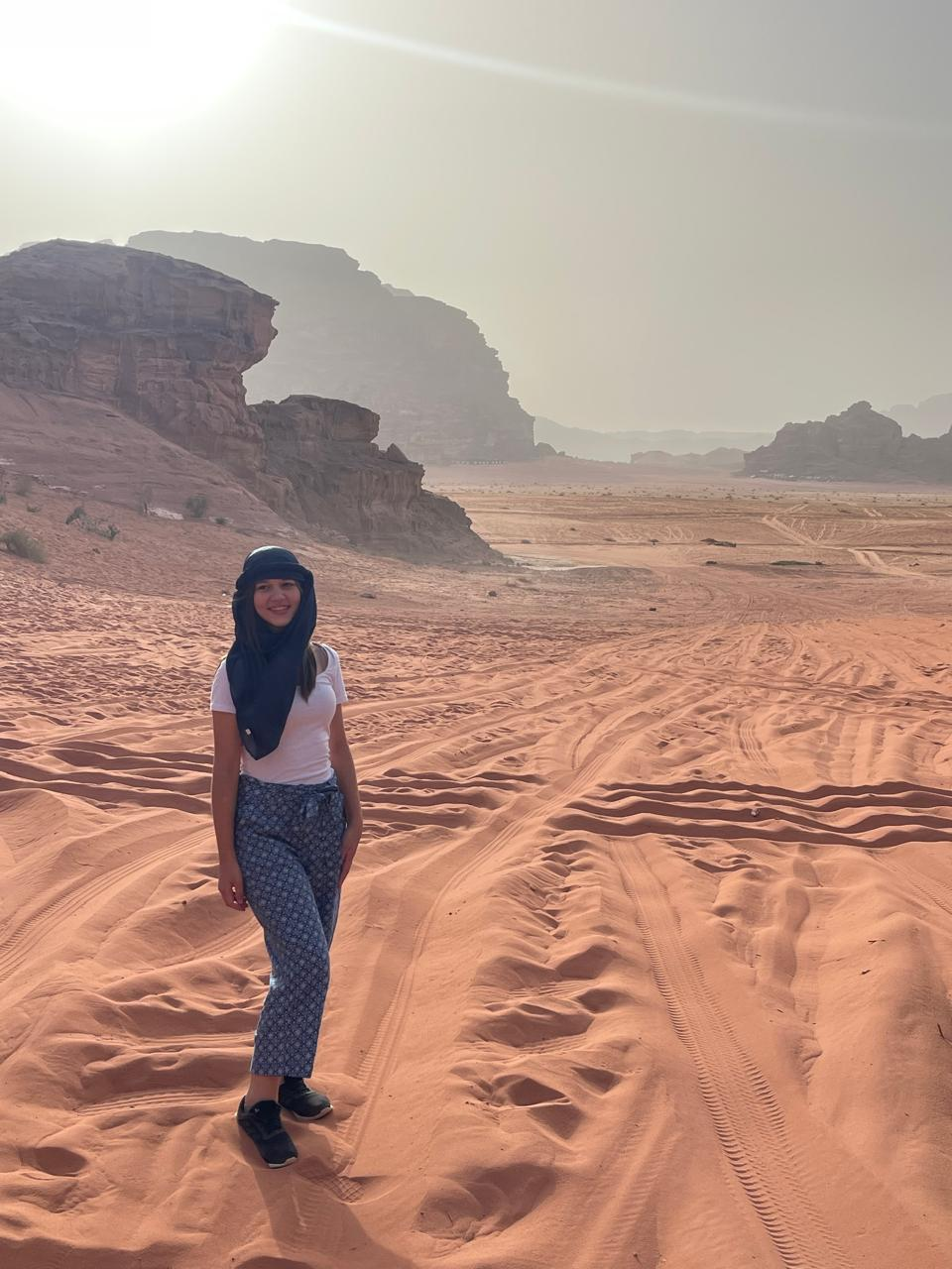 A person in a white shirt and blue patterned pants stands on sunlit red desert sand with tracks, wearing a dark headscarf. Rocky cliffs in the background.