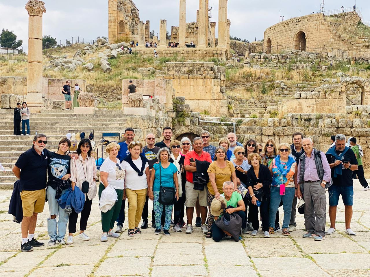 A group of tourists stands smiling in front of ancient Roman ruins, featuring towering columns and stone structures. The atmosphere is lively and cheerful.