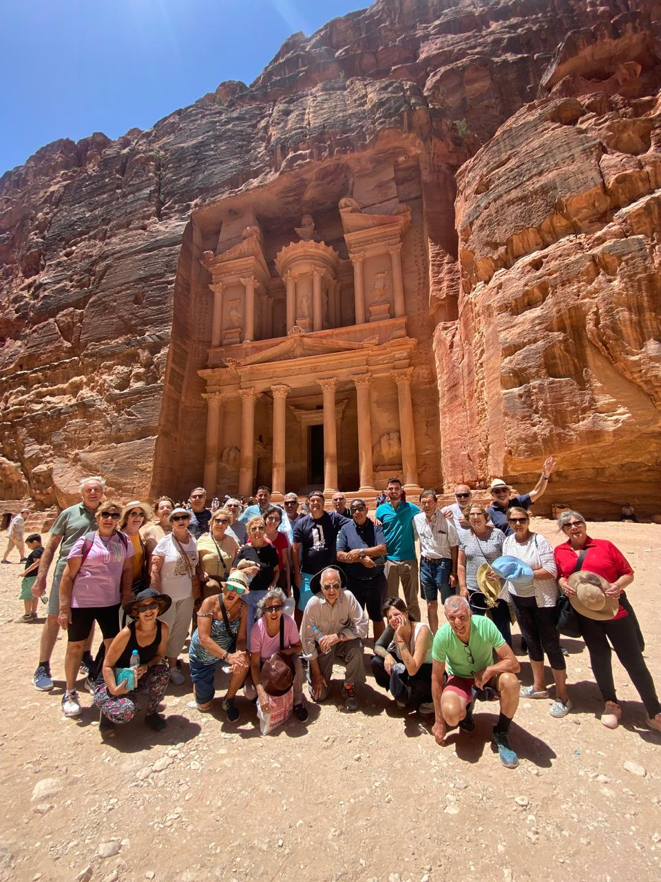 A group of people posing in front of the ancient rock-carved Al-Khazneh in Petra, Jordan. The scene is sunny, highlighting the vibrant sandstone.