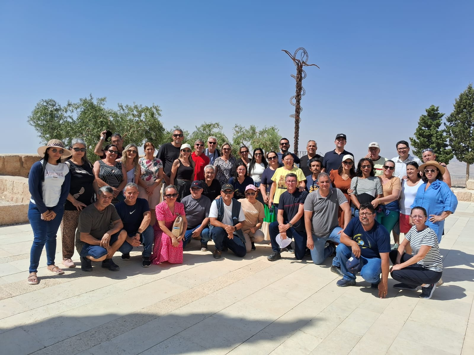 A group of around 40 tourists posing cheerfully on a sunny day at Mount Nebo in Jordan, with panoramic views of the Jordan Valley and clear skies in the background.