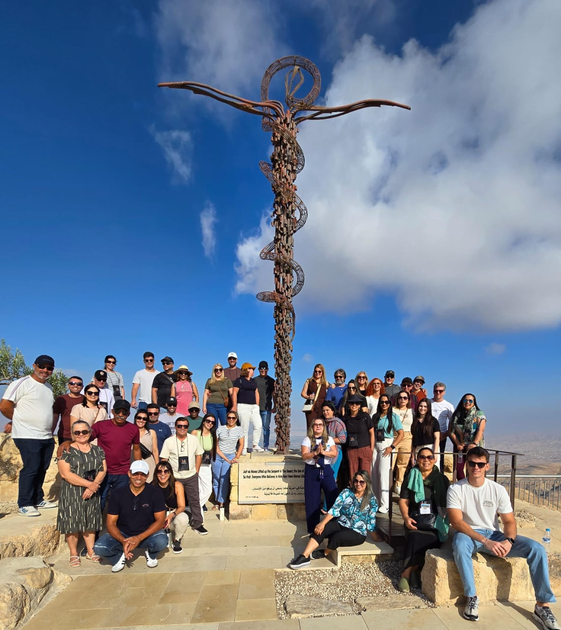 A large group of tourists posing in front of the Brazen Serpent Monument at Mount Nebo, Jordan, with the twisted serpent sculpture and clear blue sky in the background.