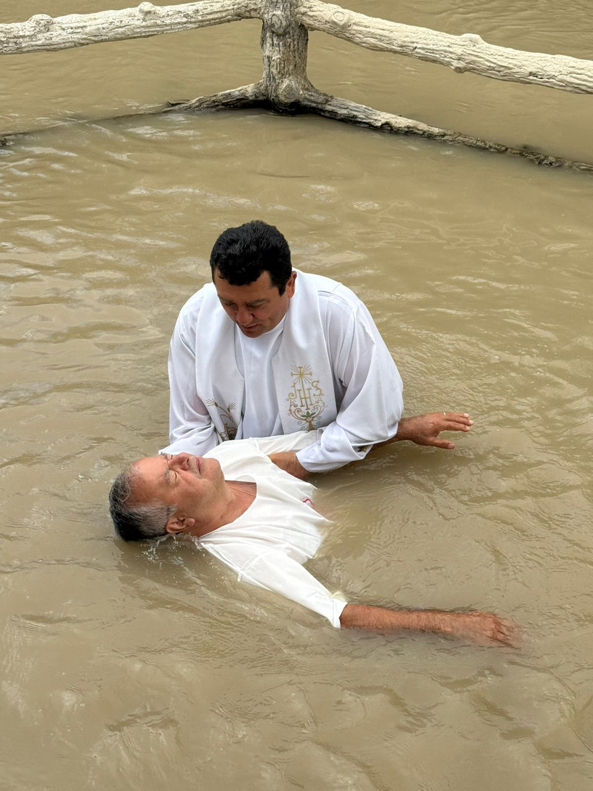 A man in a white robe performs a baptism in Jordan River, gently lowering an older man wearing a white shirt. A wooden barrier is visible in the background.