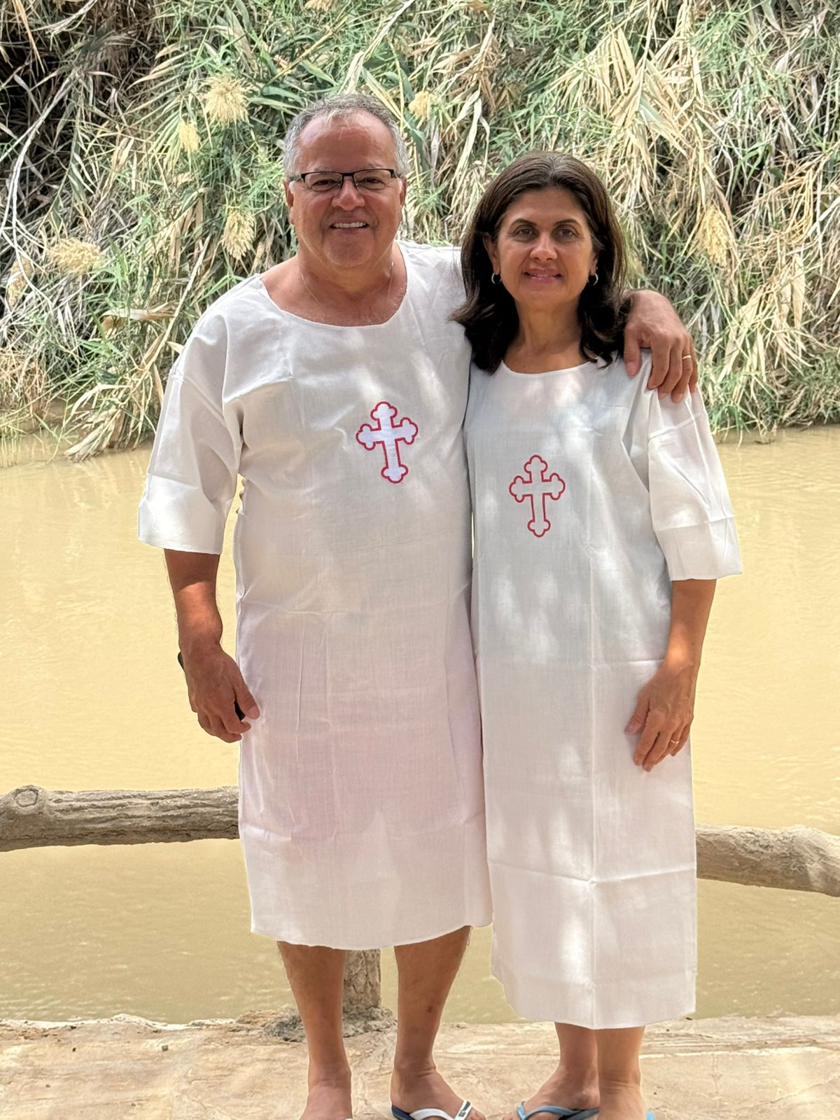 A man and woman stand side by side in front of Jordan River at the baptism site, wearing white garments with red crosses. They smile, conveying a peaceful and joyous mood.