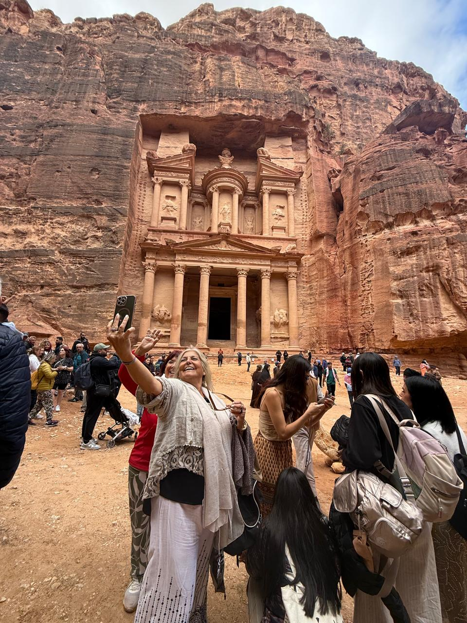A group of tourists, with one woman taking a selfie, stand in front of the iconic rock-carved facade of Petra's Treasury in Jordan. The scene is lively and vibrant.