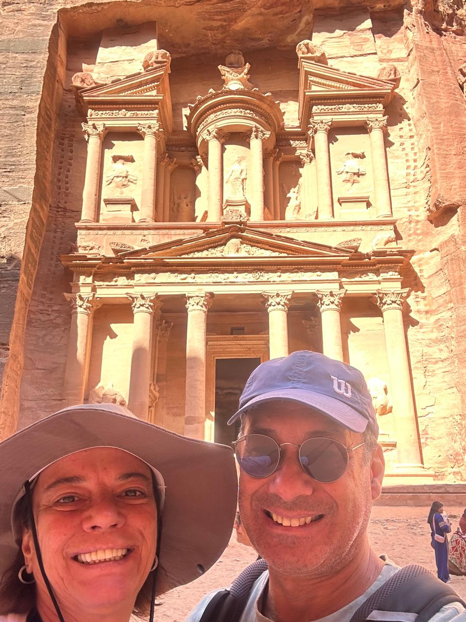 A smiling couple in hats and sunglasses poses in front of Petra's ancient stone-carved facade glowing in bright sunlight, conveying joy and adventure.