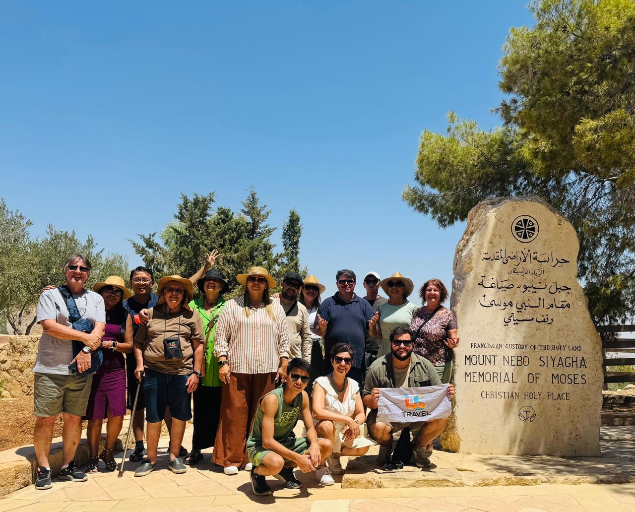 A group of tourists in casual attire pose cheerfully at Mount Nebo, near a stone marker reading "Memorial of Moses." Blue sky and trees create a serene backdrop.