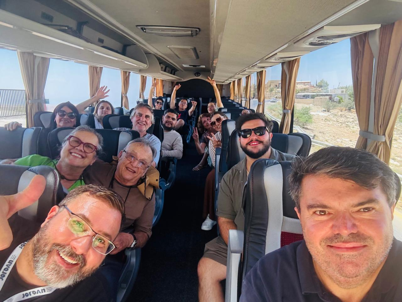 Group of cheerful people on a bus, smiling and waving at the camera. The bright interior creates an upbeat, friendly atmosphere.