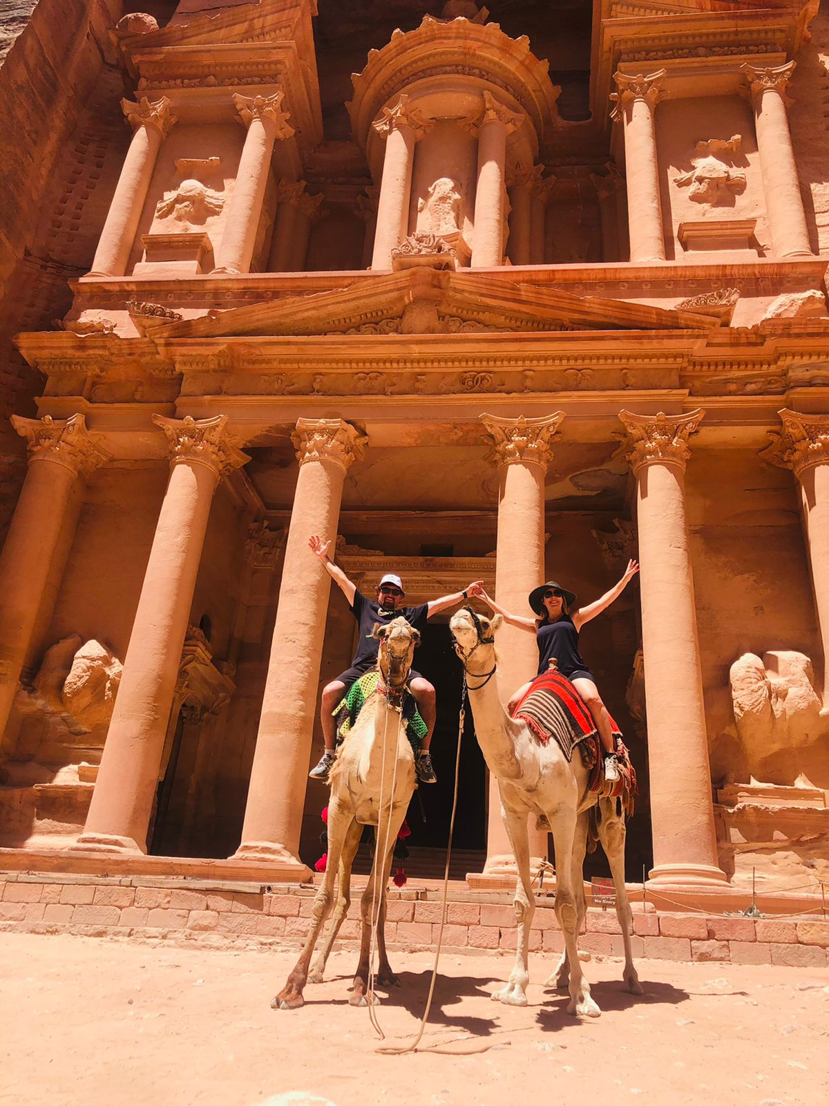 People sit on camels with arms raised in front of Petra's Treasury. The ancient structure features grand columns, exuding a sense of adventure and history.
