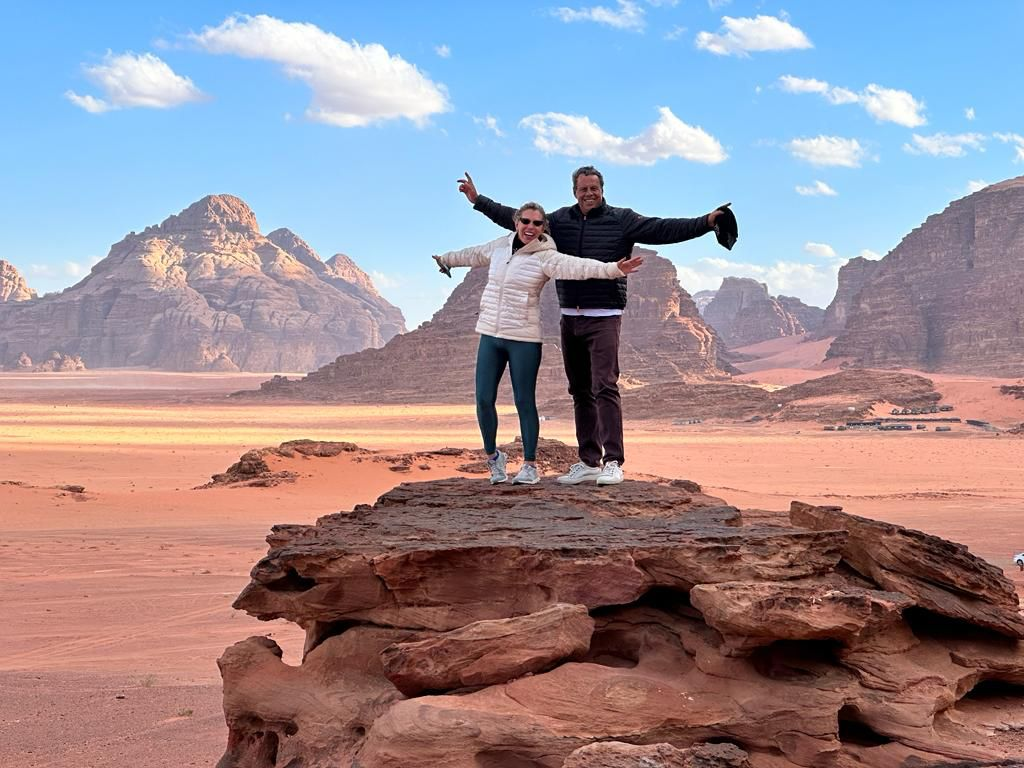 Couple joyfully poses with arms outstretched on a rocky ledge in Wadi Rum desert , framed by vast red sand and majestic mountains under a blue sky.